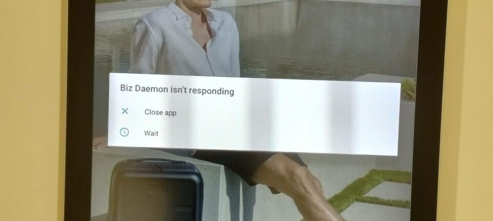 Electronic sign with pop-up text 'Biz Daemon is not responding' over an image of a man in shorts sitting on a wall with a wheeled suitcase
