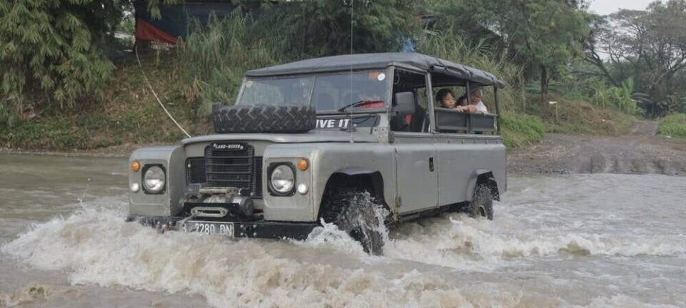 A convoy of classic Land Rover vehicles, also known as Landy, crosses a shallow river in a tropical outdoor setting,
