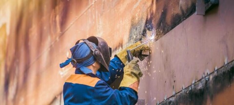 Welder on a scaffold performing maintenance on the rusty hull of a massive vessel. Pic by: Deniss Dronin / Shutterstock