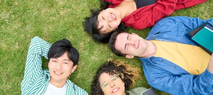 Four young people lie on green grass in colorful clothing, viewed from above, smiling and relaxing.
