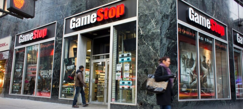GameStop storefront with large windows and red signs on a street corner, with pedestrians walking past.