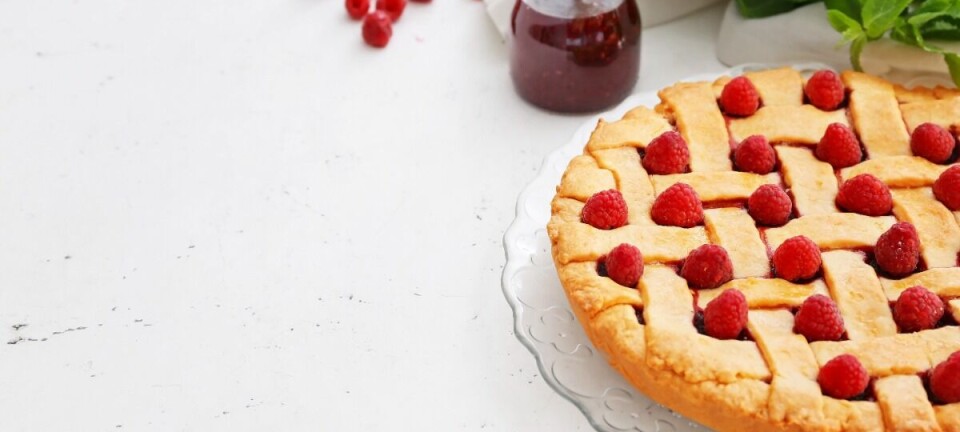 A raspberry tart on a glass plate with a jar of jam, red berries, and green leaves on a white surface.